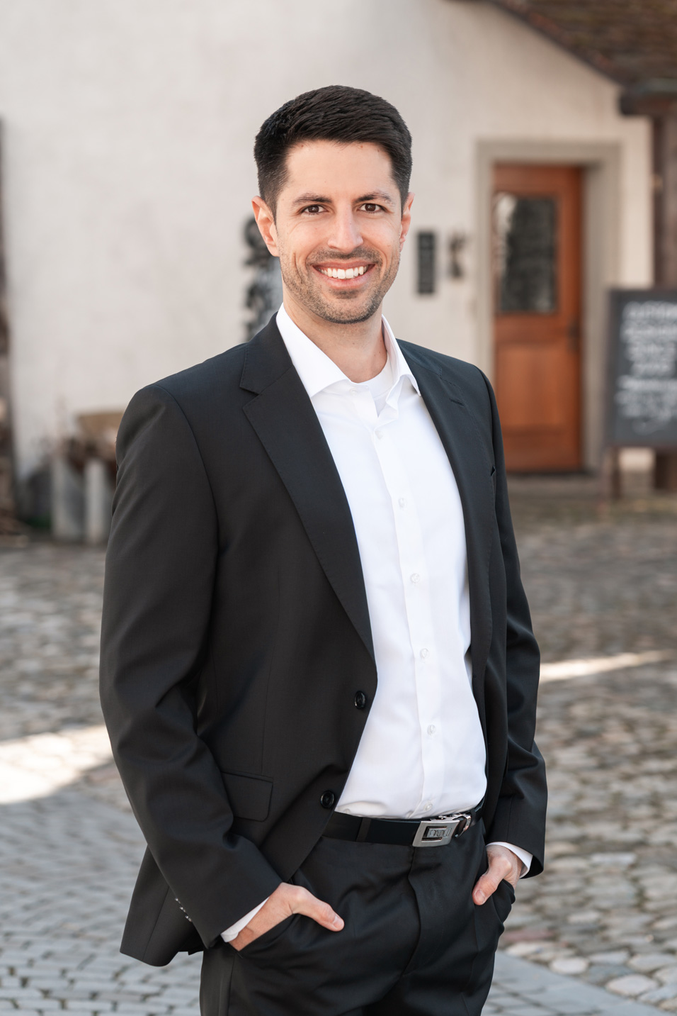 Pascal Droux lacht in formeller Kleidung Pascal Droux trägt einen schwarzen Anzug und ein weisses Hemd, während er auf einem gepflasterten Platz vor einem historischen Gebäude lächelt. Der Hintergrund zeigt eine rustikale, aber elegante Architektur, die zu seinem professionellen Erscheinungsbild passt.