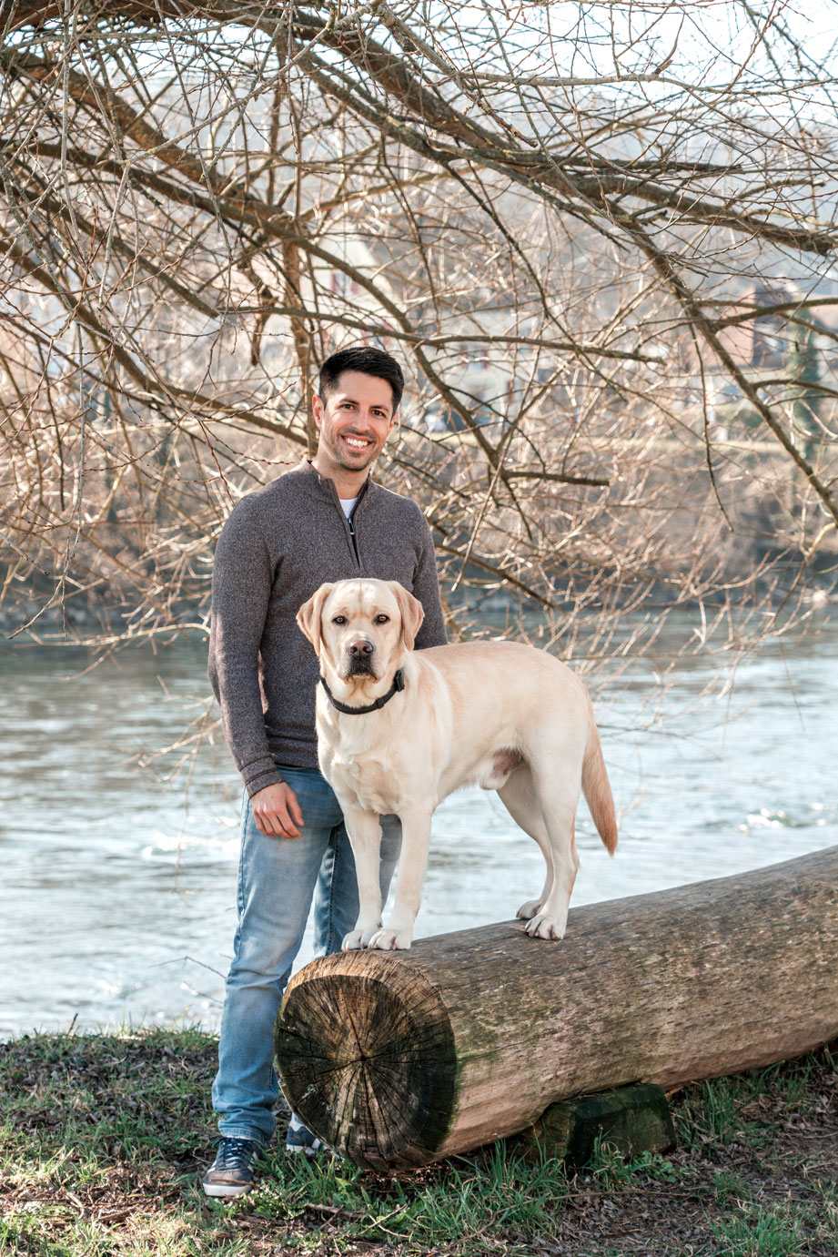 Pascal Droux mit seinem Hund am Flussufer Pascal Droux steht mit einem Lächeln am Ufer eines Flusses, während sein Hund, ein Labrador, auf einem Baumstamm steht. Der Hintergrund zeigt einen ruhigen Fluss und Bäume ohne Blätter, was die frische Atmosphäre des Frühlings vermittelt.