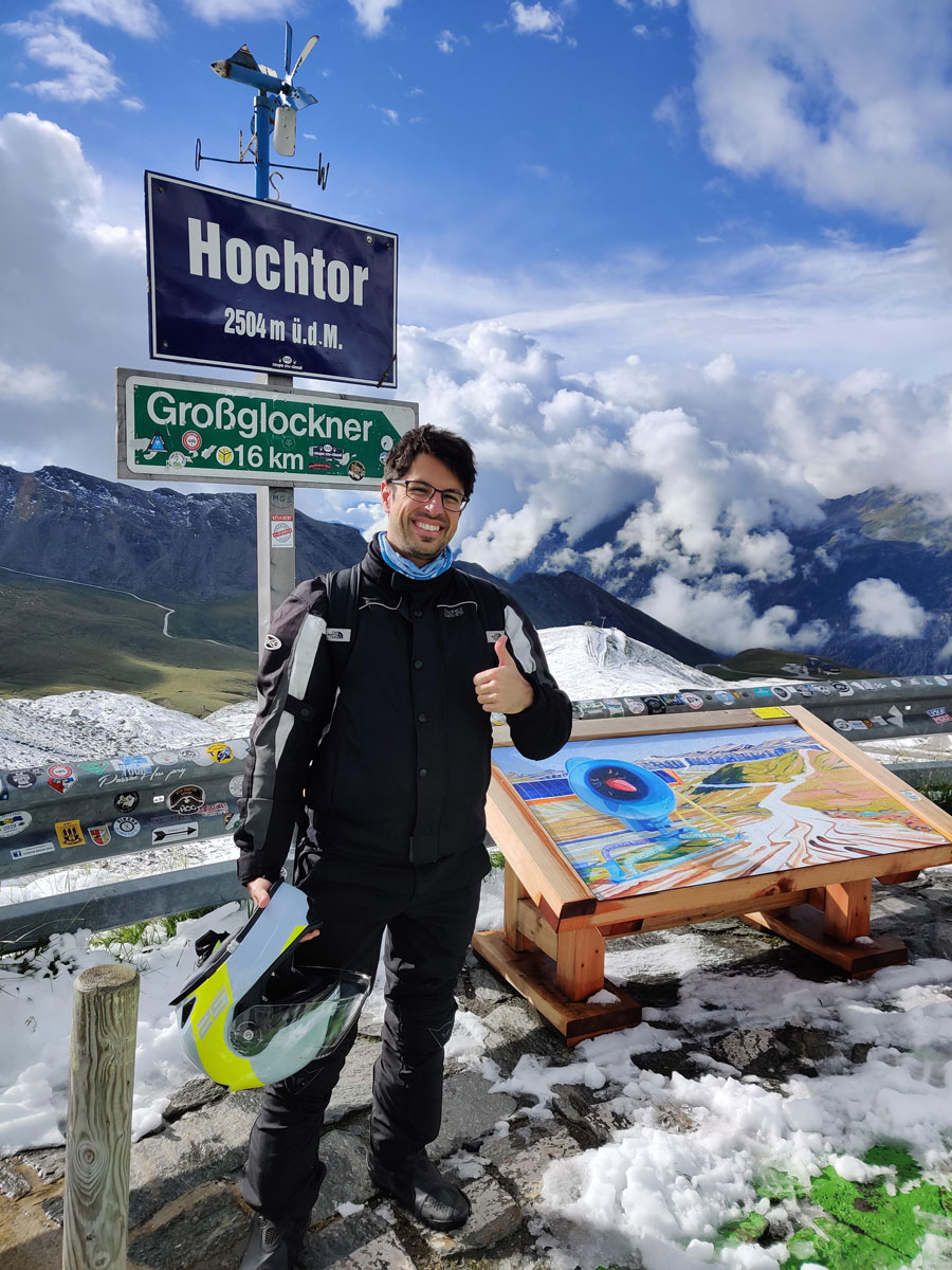 Pascal Droux auf dem Hochalpenpass Hochtor Pascal Droux steht auf dem Hochtor Pass (2.504 m ü.d.M.) mit einem Daumen hoch, während er einen Motorradhelm in der Hand hält. Im Hintergrund ist das beeindruckende Bergpanorama der Alpen zu sehen, mit Wolken und Schnee. Ein Informationsschild zeigt die Entfernung zum Grossglockner (16 km) an.